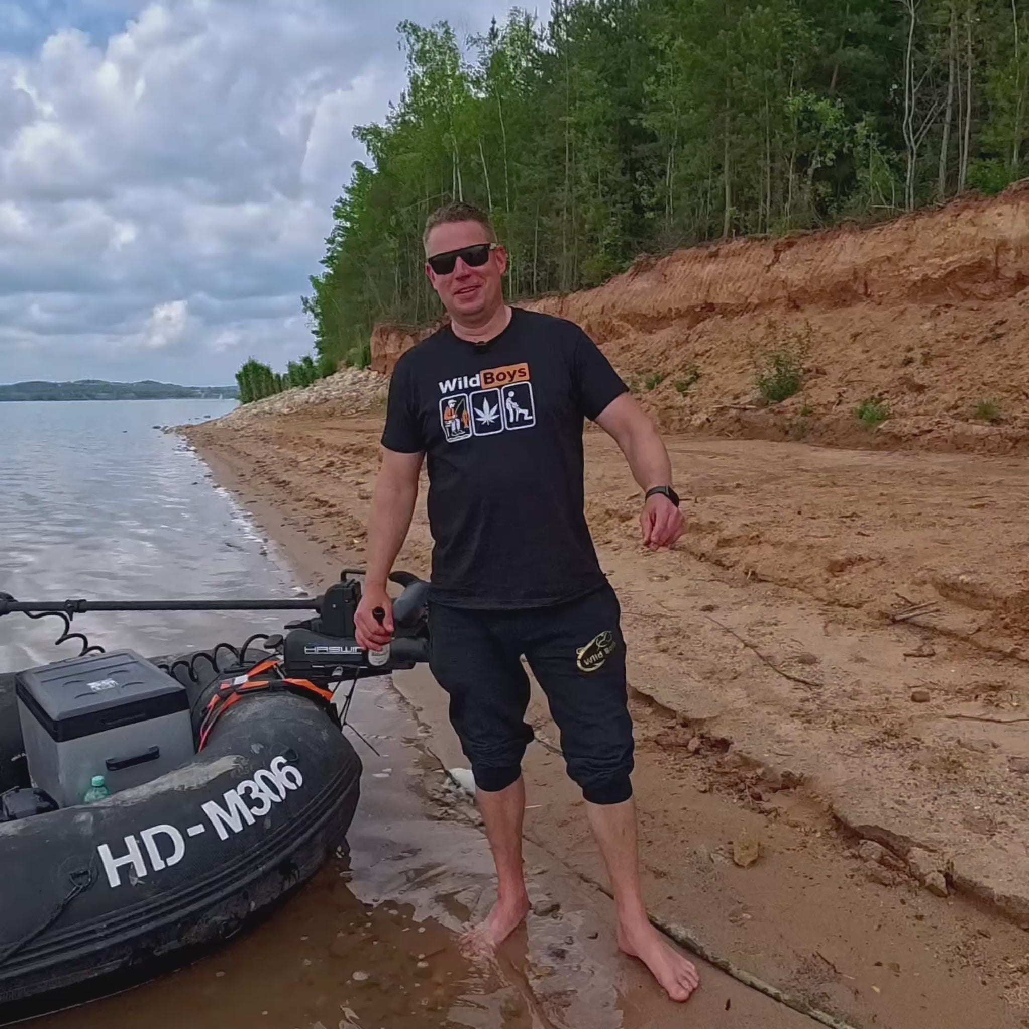 Video eines Mannes am Seeufer, der ein Wild Boys Angler T-Shirt trägt und neben einem Schlauchboot steht. Outdoor-Szene mit Wasser, Sandstrand und Wald im Hintergrund.